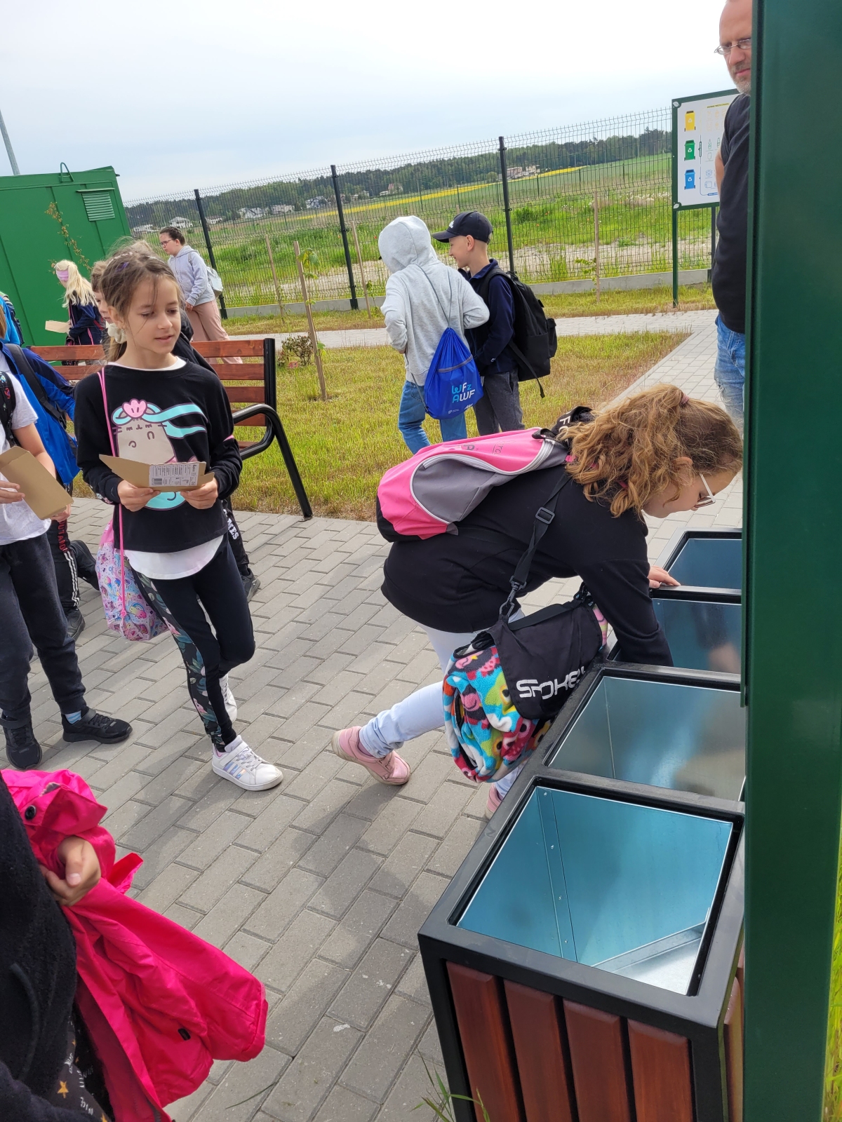 Dziewczynka z różowym plecakiem wrzuca przedmiot do odpowiedniego pojemnika na odpady.